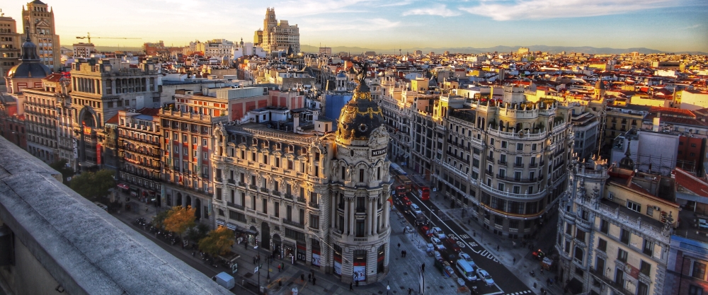 Aerial view of Madrid's Gran Vía, highlighting the black and gold dome of the Metropolis Building at the intersection, with the city skyline stretching out beneath a clear sky.